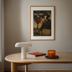 Framed poster of a cow in a suit with a pipe on a wall above a wooden table with a lamp, book, and candle.