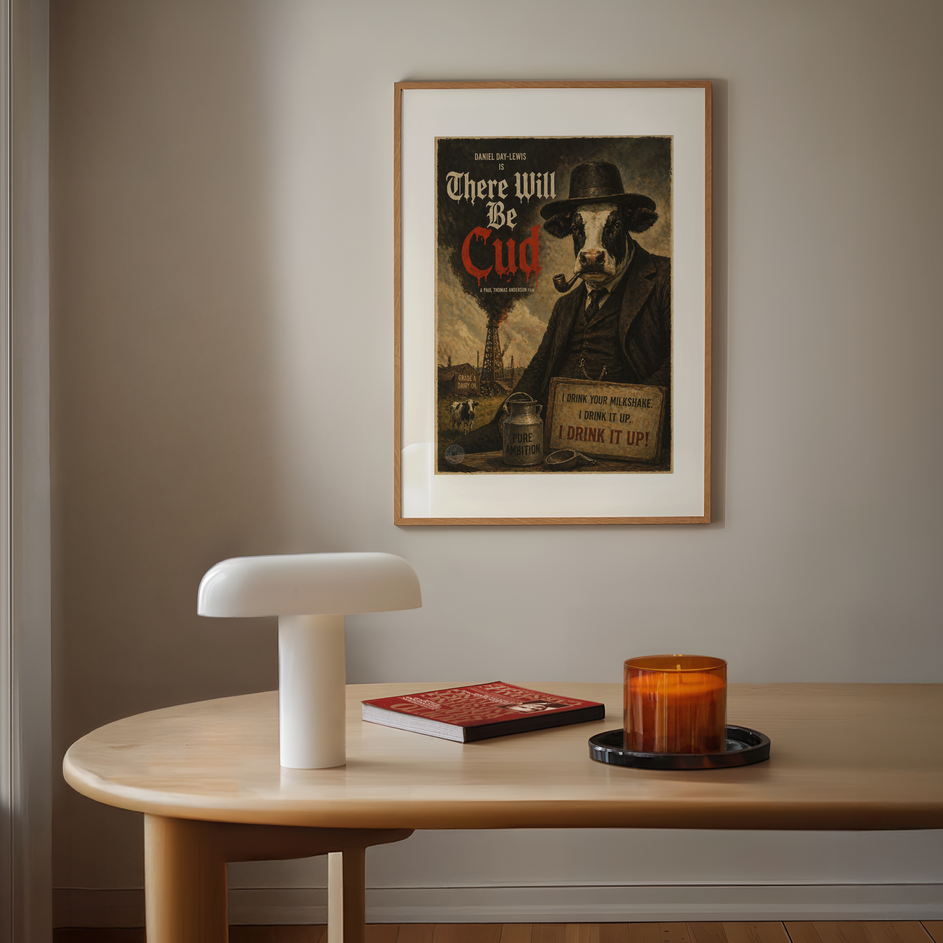 Framed poster of a cow in a suit with a pipe on a wall above a wooden table with a lamp, book, and candle.