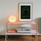 Pink shelf with a lamp, books, and a vintage radio player against a white wall with a framed inspector gadget matrix poster.