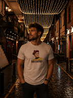 A man wearing a light gray graphic t-shirt with a portrait of Tam O'Shater, standing in a narrow, wet cobblestone alleyway at night under warm hanging string lights.