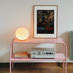Pink shelf with a lamp, books, and a framed waterboy vs hellboy poster on a white wall.