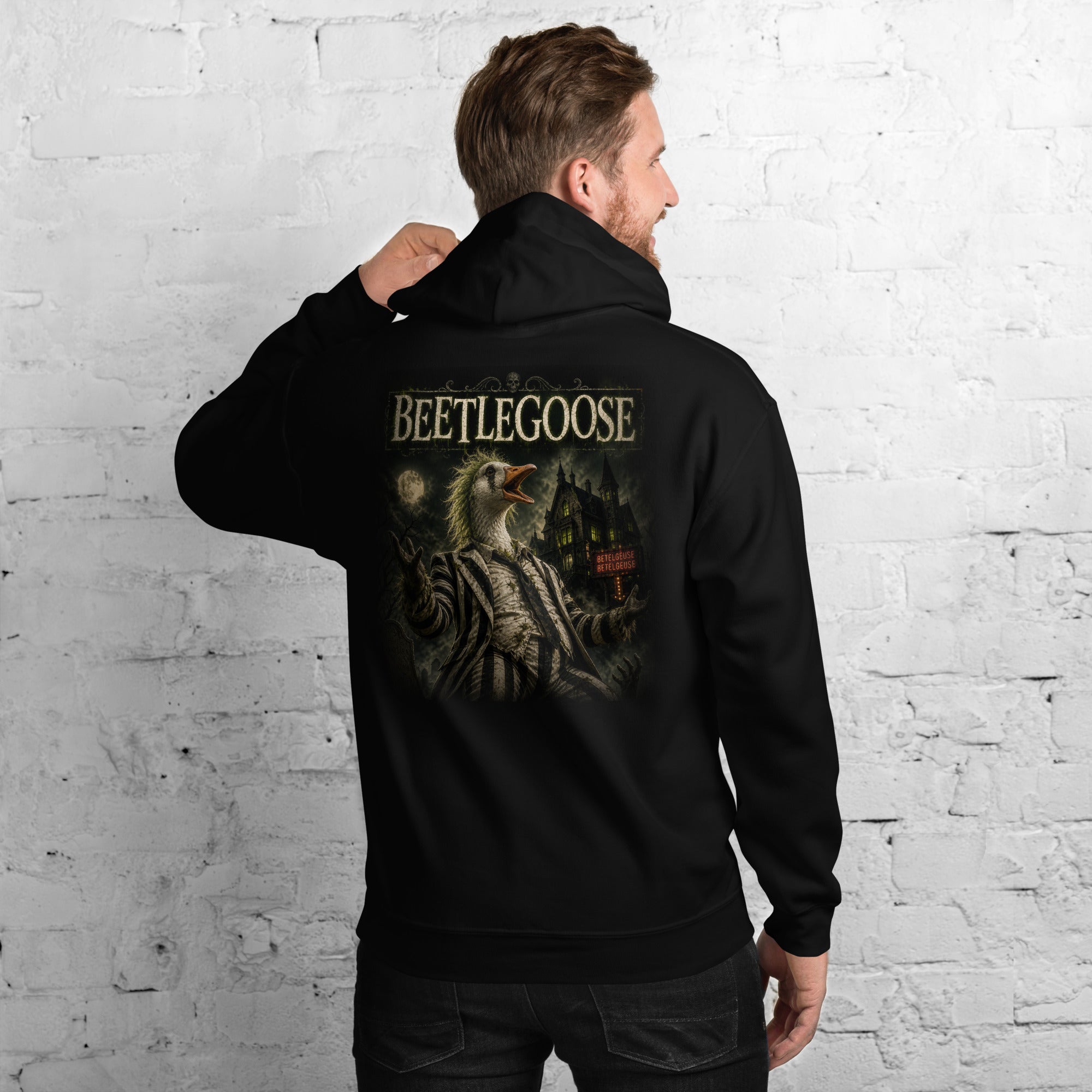 Man wearing a brown hoodie with a graphic and text beetlejuice on the back, against a white brick wall.