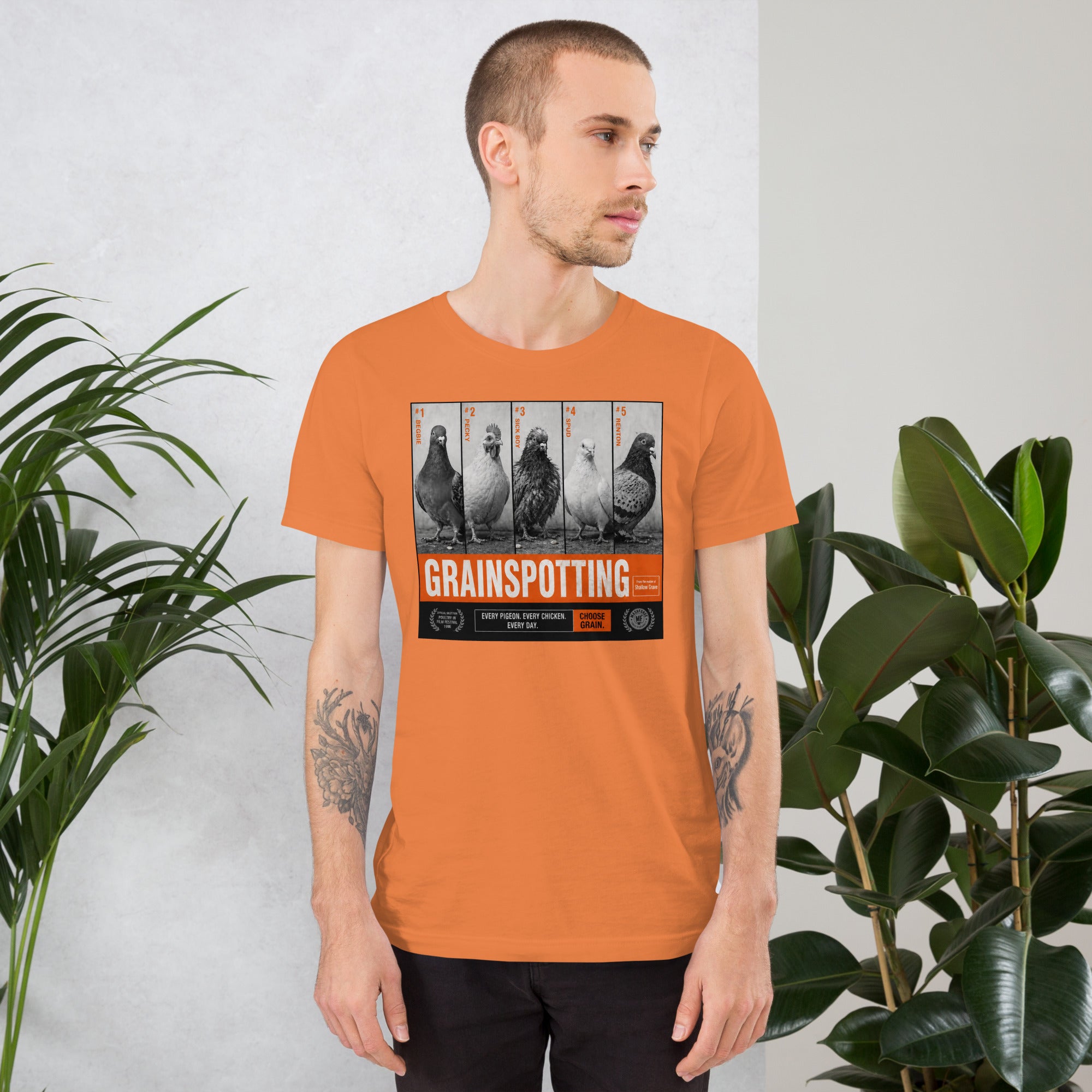Man wearing an orange t-shirt with a graphic design and text, standing against a light gray background with plants.