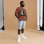 Man wearing a brown t-shirt with a funny teletubby style graphic and light blue shorts standing against a beige backdrop.