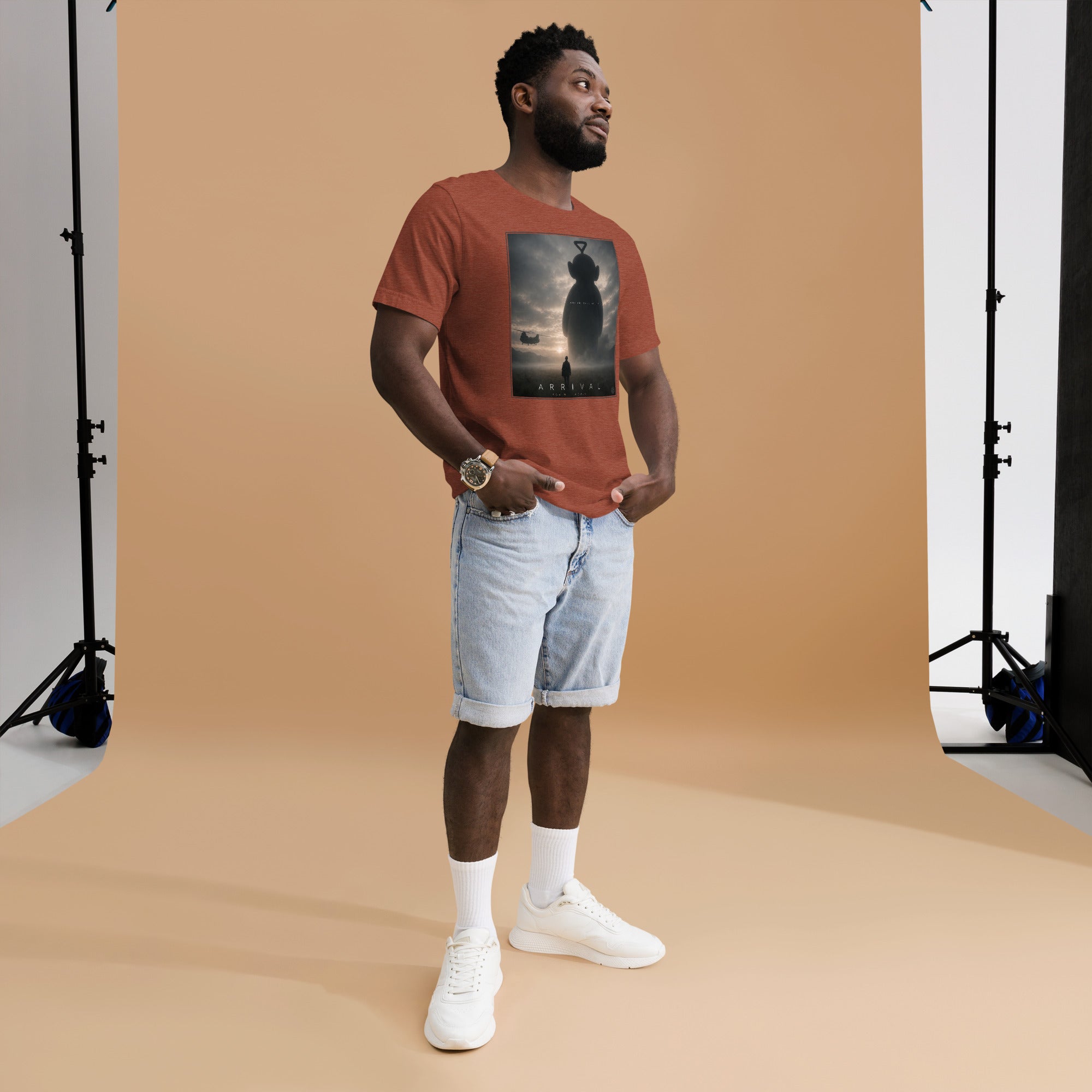 Man wearing a brown t-shirt with a funny teletubby style graphic and light blue shorts standing against a beige backdrop.