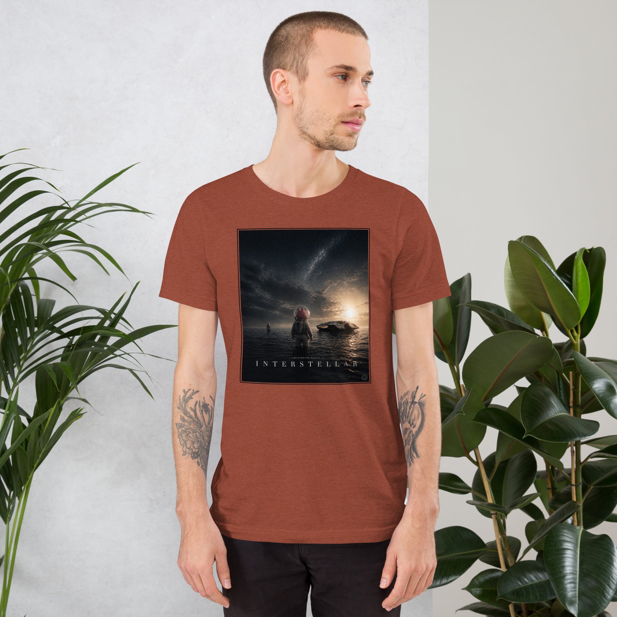 Man wearing a brown t-shirt with 'Interstellar' graphic design, standing between two plants.