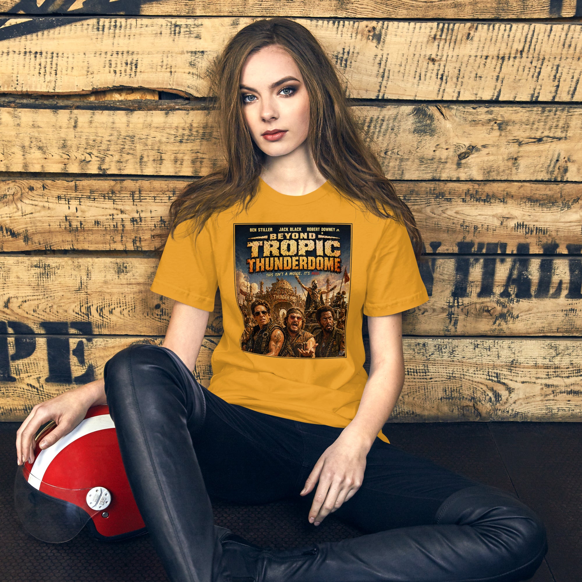 Woman wearing a yellow t-shirt with Beyond Tropic Thunderdome design, sitting against a wooden wall.