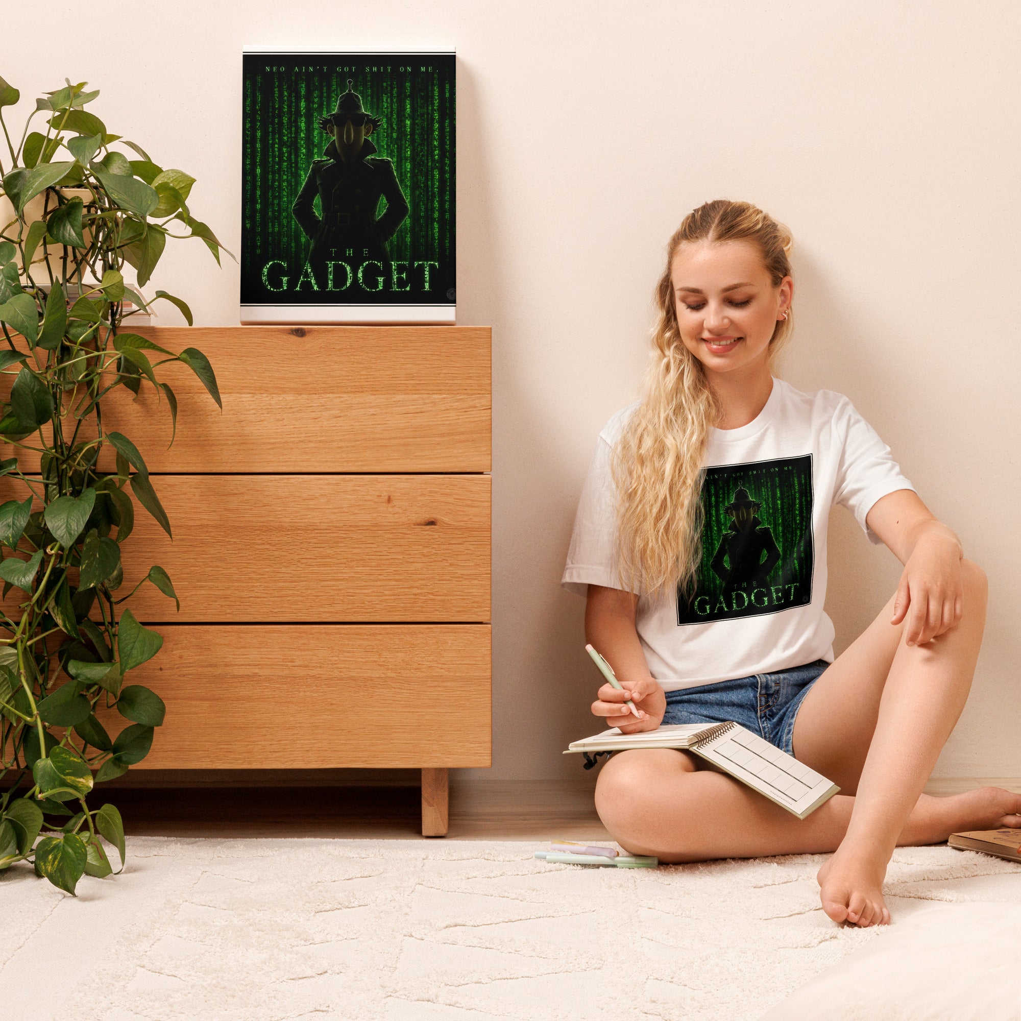Woman sitting on the floor with a book, wearing a white t-shirt with a graphic design, in a room with a wooden dresser and plant.