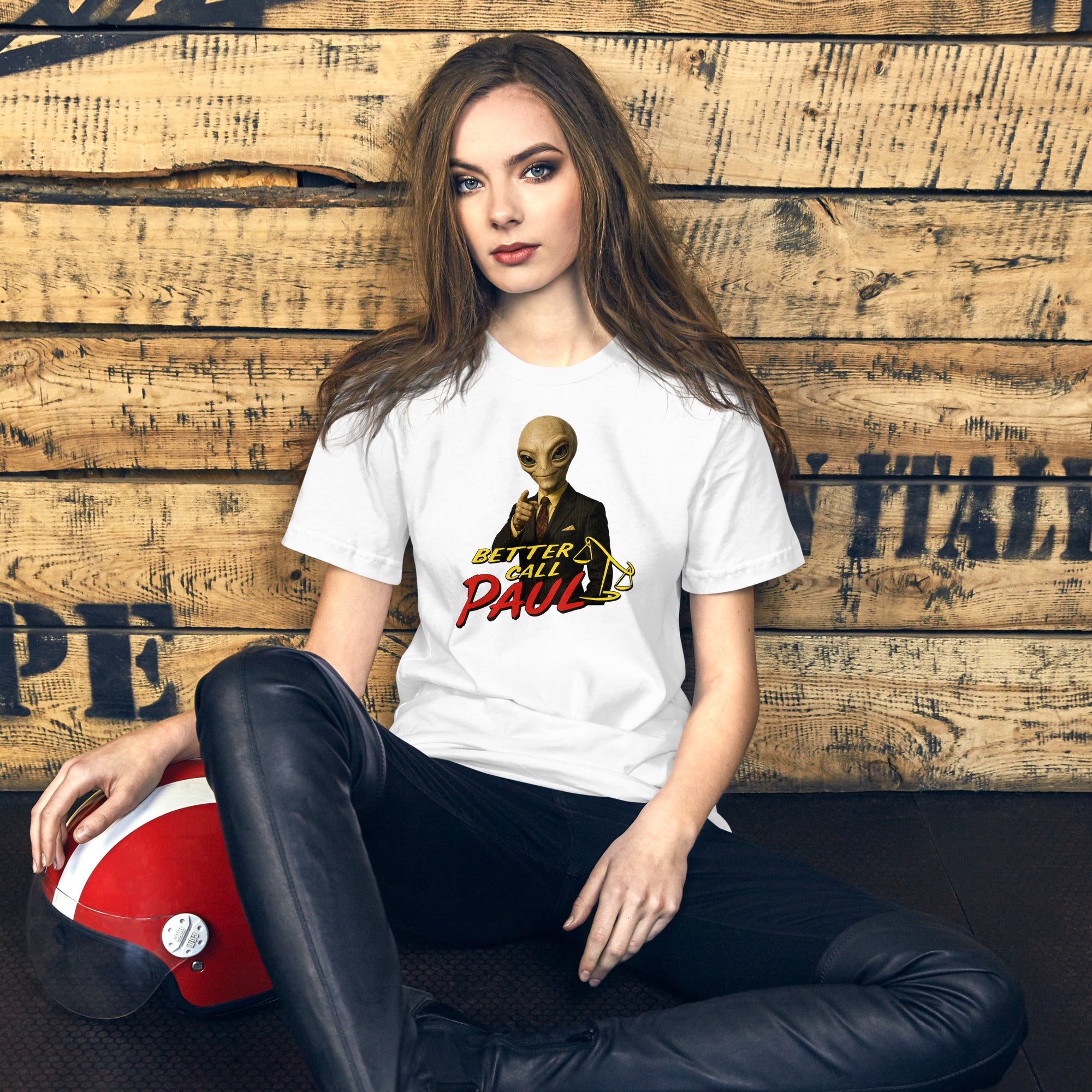 A woman sits cross-legged against a rustic wooden wall, wearing a white graphic T-shirt with a alien character and the text 'Better Call Paul' in red and yellow, paired with black pants. A red and white helmet rests beside her.