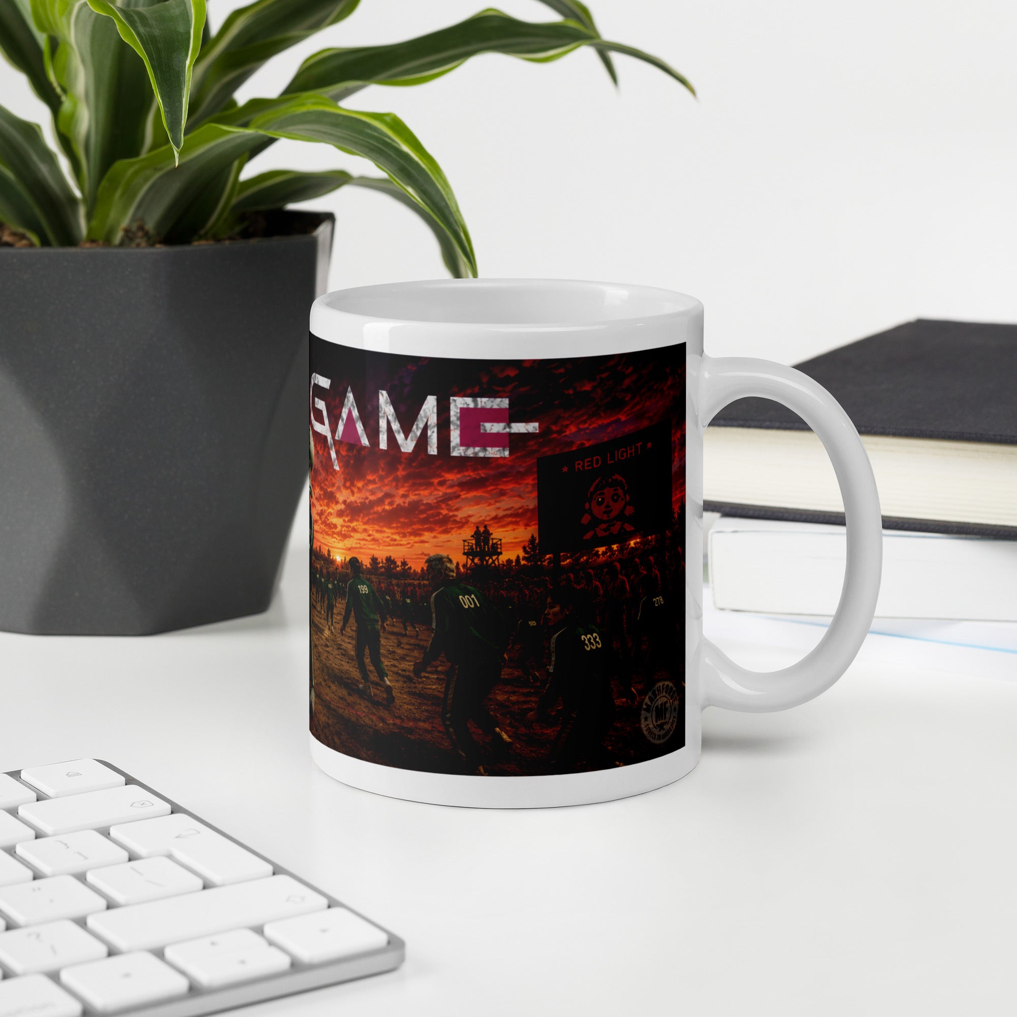Mug with a sunset design and 'squid  GAME' text on a desk with a plant and books.